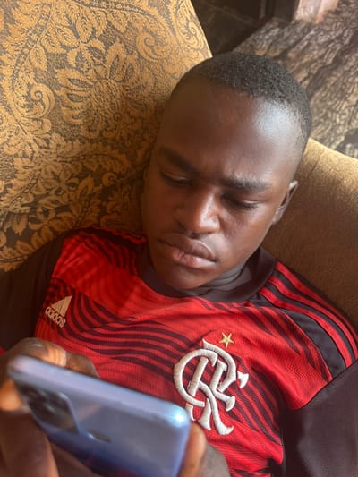 An individual is depicted in Umuoye, Nigeria, seated or reclining on a brown fabric surface with a gold damask pattern. The subject is a male with dark skin and short dark hair, viewed from above, looking downward with a focused expression. He is wearing a red and black striped short-sleeved shirt featuring the white Adidas logo on the left chest and a white embroidered emblem with a golden star above it on the right chest, consistent with the Club de Regatas do Flamengo crest. Both hands are holding a light-colored smartphone, possibly light blue or purple, which is partially visible at the bottom of the frame, emitting light from its screen. The overall lighting appears bright, highlighting the subject's forehead and the phone.