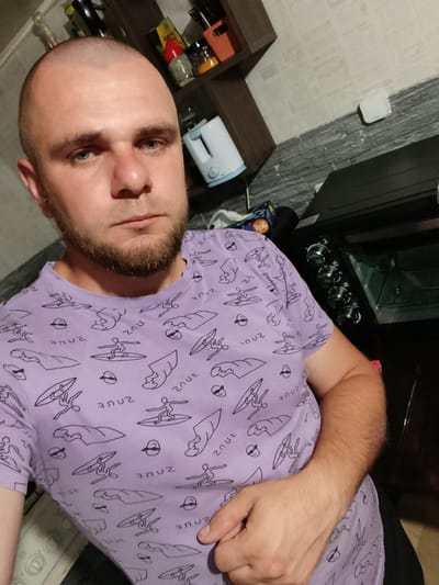 A bald man with a short beard is taking a selfie in what appears to be a kitchen. He is wearing a purple t-shirt with a repeating pattern of surfers. In the background, there is a shelf with various items, a white electric kettle, and a black oven. The walls appear to be tiled. The specific location in Pishchane, Ukraine, is not discernible from the image.