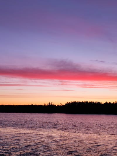The image depicts a sunset over water, likely a lake or river, with a silhouette of trees along the horizon. The sky is layered with colors transitioning from pink and orange near the horizon to purple and blue towards the top. The water reflects the colors of the sky, creating ripples and patterns on the surface. The scene is likely taken near Vasil'evskoe, Russia, based on the prompt, suggesting this may be a natural setting near the city, though without additional information, the specific location remains speculative. There are no people visible in the scene. The dominant subjects are the sky, water, and trees. The interaction is the reflection of the sky in the water. The environment is a natural setting, likely at twilight.