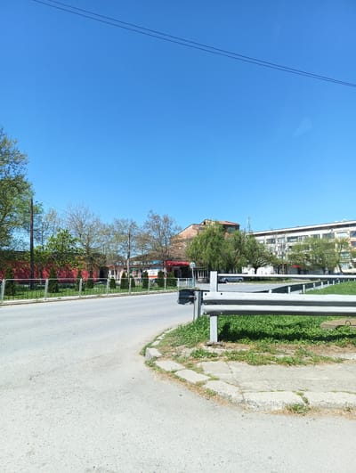 The image shows an urban street scene in Straldzha, Bulgaria, on a sunny day. The sky is clear and blue. The street pavement is light grey. A metal guardrail runs along a grassy area to the right of the street. Across the street, there's a red building with star decorations on its facade, partially obscured by trees. Further down the street, other buildings are visible, including a white building with multiple windows. A car is parked near the white building. Electrical wires stretch across the sky.