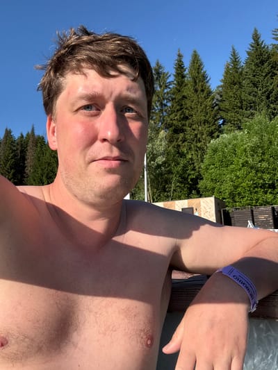 A shirtless man with brown hair and blue eyes takes a selfie outdoors. He has a light beard and is wearing a purple wristband. The background shows a clear blue sky, tall evergreen trees, and some buildings. The setting appears to be a park or resort in Nechkino, Russia, given the text on the wristband which translates to "Nechkino". The lighting suggests it is daytime, with a shadow cast across the man's chest.