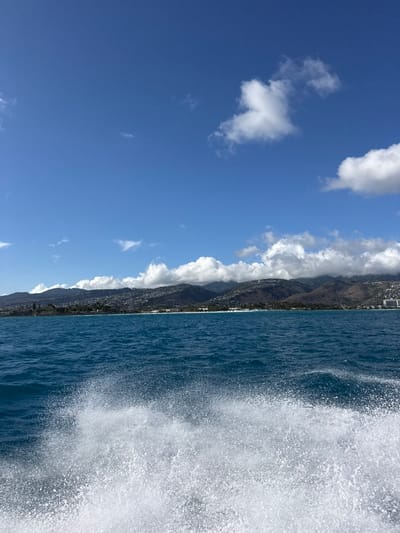 The image displays a clear daytime view of the coastline of East Honolulu, Hawaii, United States, as observed from the water. In the immediate foreground, a significant volume of white, turbulent water and spray is present, indicative of a moving vessel's wake. Beyond the wake, the midground is dominated by the calm, deep blue-green waters of the Pacific Ocean. The background features a continuous landmass comprising multiple green, tree-covered mountains and hills, upon which numerous residential structures are visible, extending up the slopes. A narrow white sand beach lines the base of the mountains along the coast. The sky occupies the upper portion of the frame, a clear blue with scattered white cumulus clouds; some clouds are positioned above or partially obscuring the mountain peaks. No individuals or specific actions beyond the wake generation are identifiable.