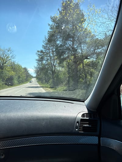 The image depicts a view from inside a vehicle traveling on a road. The exterior scenery, visible through the passenger-side window, shows a winding road lined with trees under a clear, blue sky. The vehicle's dashboard is partially visible on the right side of the frame, featuring an air vent and textured material. The precise location within the city of Levski, Bulgaria, cannot be determined from the visual information provided. The image does not show any people.