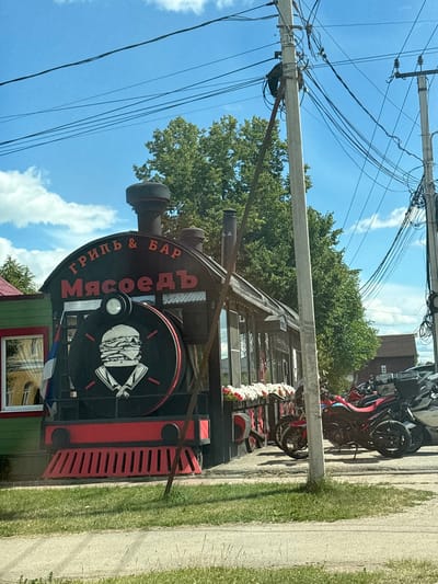 The image depicts the exterior of a restaurant designed to resemble a train. The restaurant, identified by the Cyrillic text "ГРИЛЬ & БАР Мясоедъ" (Grill & Bar Myasoyed) on its facade, is located in the city of Pereslavl-Zalessky, Russia. The front of the building is stylized as the engine of a steam locomotive, complete with a circular emblem featuring a stylized burger and two crossed knives. Red decorative elements, resembling the train's wheels and grill, are visible at the base. The restaurant is flanked by a green building on the left, and a row of motorcycles and a car are parked in front of it on the right. The scene is set against a backdrop of trees and a blue sky with scattered clouds. Electrical poles and overhead wires are present in the foreground and midground. The overall impression is of a unique, themed dining establishment in a suburban or roadside setting.