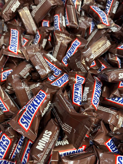 A close-up, overhead shot shows a large pile of unwrapped "Snickers Minis" chocolate bars in their individual brown wrappers. The wrappers feature the distinctive red, white, and blue "SNICKERS" logo, often with "MINIS" written in white text below it. The bars are densely packed, obscuring much of the background. The scene appears to be a retail display or storage area in a store or warehouse. The location is identified as Sokilnyky, Ukraine.