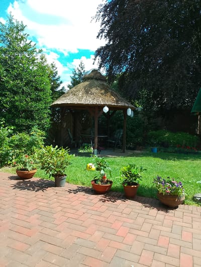 The image shows a well-maintained garden with a thatched-roof gazebo as the central feature. The gazebo is made of wood and has a circular design, with tables and chairs visible inside. In front of the gazebo, a lush green lawn extends towards a brick patio. Several potted plants of varying sizes and types are arranged on the patio, including some with colorful flowers. Behind the gazebo, dense foliage and trees create a natural backdrop. The sky is bright blue with fluffy white clouds, indicating a sunny day. The overall impression is one of a peaceful and inviting outdoor space, likely a private garden in Rheda-Wiedenbrück, Germany, given the context. There are no people visible in the image.
