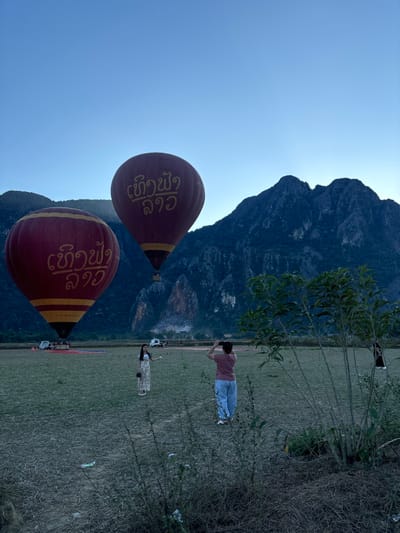 The scene is set in a grassy field in Hong Ngern Village, Laos, under a clear, light blue sky at what appears to be dawn or dusk. Two large, red hot air balloons, each bearing yellow Lao script (reads "ເທີງຟ້າ ລາວ"), are central to the image. The left balloon is fully inflated and grounded, with several white utility vehicles visible behind it on the field. The right balloon is also fully inflated and slightly airborne, ascending above the ground. In the midground, two individuals are present. A female subject, wearing a light-colored patterned dress, stands facing the right, with her right arm slightly extended, posing for a photograph. To her right, a second subject, viewed from behind, holds a dark handheld device, actively photographing the female subject with the hot air balloons and mountains in the background. The background is dominated by steep, dark, forested karstic mountains. Sparse green vegetation and dry grass cover the immediate foreground.