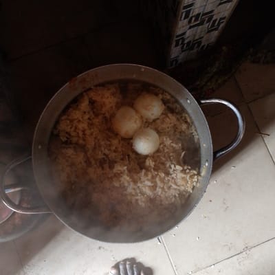 The image shows a large metal pot containing rice and three boiled eggs. The pot is sitting on a tiled floor, with a portion of a foot visible at the bottom of the image. Steam is rising from the pot, indicating the contents are hot. The background shows a portion of a wall and some objects, likely in a kitchen or dining area. The image suggests a meal being prepared or served in a home or residential setting in Adanisievbe, Nigeria.