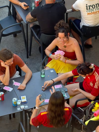 This image shows a group of people playing cards at an outdoor cafe table in Las Rozas de Madrid, Spain. The scene appears to be during the daytime, with ample natural light. The people are dressed casually, with some wearing t-shirts and shorts, suggesting warm weather. The cafe tables and chairs, along with the stone-like ground, indicate an urban or public outdoor setting.
The central activity is a card game, with several hands of pink-backed cards visible on the tables. One person is holding a fan of cards, while others are reaching for them or looking at their hands. There are also drinks on the table, including a glass of water and a can of what appears to be a soft drink. A smartphone is also present.
Notable details include the tattoos on the arms of one of the individuals, the various bracelets worn by some of the players, and the use of QR codes on stickers on the table. One person is wearing a red shirt with yellow stripes, reminiscent of a sports jersey. The expressions of the people are not clearly discernible due to the overhead angle, but the relaxed posture suggests an informal and enjoyable gathering.
Visible text includes "iron cafe motors" on a white t-shirt in the upper left corner. Another partially visible text on a sticker on the table reads "AMB". There is also text on the can which appears to say "Nestle" and "Blue Sky".