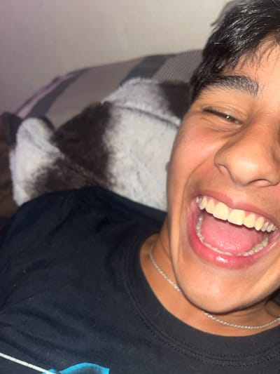 A close-up image features a person smiling widely with their mouth open, revealing their teeth and tongue. The person wears a dark t-shirt and a thin silver necklace. The background consists of a patterned cushion or blanket in shades of brown and gray. The image is slightly tilted, giving a candid and informal feel. The person's face is well-lit, highlighting their features and expression.