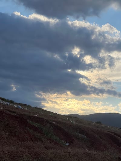 The image depicts a sloping field littered with trash in Ait Mazigh, Morocco. The foreground consists of reddish-brown soil interspersed with dry vegetation and scattered white plastic bags. In the midground, a dark, undefined mountain range is visible against a sky filled with layered clouds. The clouds range from bright white to dark gray, with the sun shining through some of the gaps.