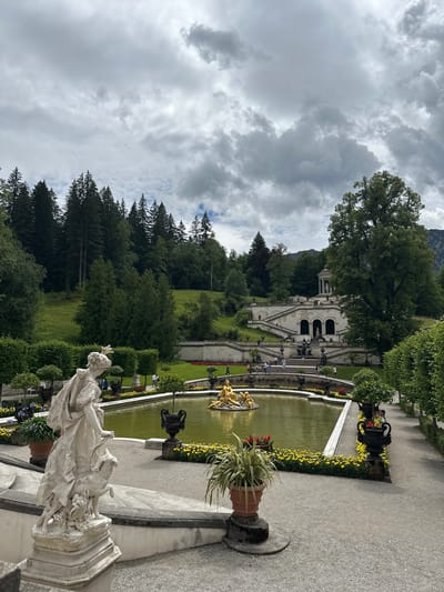 The image depicts a view of the Linderhof Palace gardens in Ettal, Germany. In the foreground, a white marble statue of a woman with a dog stands on a tiered stone platform. The statue is positioned to the left of a rectangular pond filled with murky green water. In the center of the pond, a golden sculpture of figures is visible. The pond is bordered by a low wall and a bed of yellow flowers. Beyond the pond, a grand staircase ascends towards a white building with a dome, which is part of the palace. The staircase is adorned with statues and fountains. The gardens are meticulously maintained with manicured bushes and trees. The background features a dense forest covering a hillside, leading up to a cloudy sky. Several people are scattered throughout the scene, walking on the paths and near the staircase.