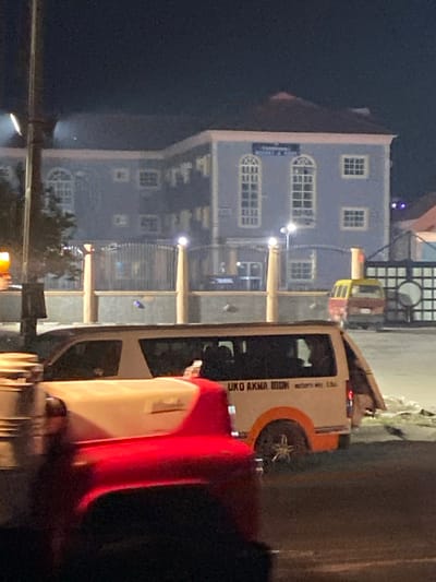 A nighttime shot captures a light blue, multi-story building with symmetrically arranged windows, some arched. Streetlights illuminate the facade. Partially visible is a parked white van with orange accents and lettering. A red vehicle dominates the foreground on the right, obscuring some of the scene. The image is slightly out of focus and appears to be taken from a moving vehicle.