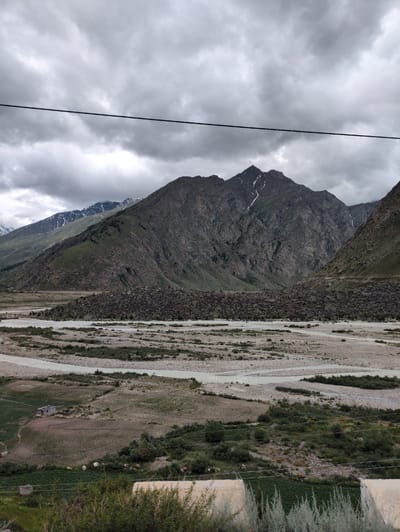 This is a vertical, eye-level shot of a mountainous landscape in Darcha, India, under a heavily overcast sky. A thin, dark power line stretches across the upper portion of the frame, bisecting the cloudy sky. The middle ground is dominated by a wide, rocky riverbed with several channels of pale, flowing water. To the left of the riverbed, in the foreground, is a small, dilapidated building with a corrugated metal roof, situated in a cultivated field. The field itself appears to be plowed in rows. Beyond the riverbed, steep, rocky mountains rise, with patches of green vegetation on their lower slopes. The peaks of these mountains are dark and jagged, with some snow visible at higher elevations. The overall mood is somber and rugged, characteristic of a remote mountain region.
