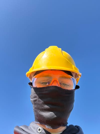 A subject is depicted in a low-angle portrait, framed from the upper chest against an unobstructed, clear blue sky, identified as being in Wellington, United Kingdom. The individual wears personal protective equipment: a yellow hard hat, orange-framed clear safety goggles over their eyes, and a black fabric face covering concealing the nose, mouth, and chin. Below the face covering, a dark grey or black hooded garment, possibly a sweatshirt or jacket, is visible, featuring a silver grommet on the left side near the neck. The subject's eyes, visible through the goggles, appear closed or significantly shadowed. The overall scene suggests an outdoor setting under bright daylight conditions.