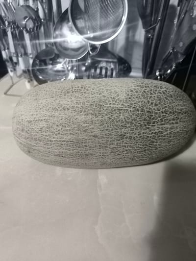 The media file shows an oval-shaped cantaloupe melon resting on a smooth, light-colored surface, possibly a countertop or table. The melon has a textured rind with a net-like pattern of light and dark lines. In the background, out of focus, is a rack holding various kitchen utensils, including ladles and what appears to be a sieve. The overall scene is dimly lit, suggesting an indoor setting, possibly a kitchen. The backdrop behind the utensils has a pattern that resembles tiles or a mural. There is no visible text in the image. The focus is entirely on the melon and the hint of kitchen items in the background, creating a still life composition. The context provided indicates the location is Kokand, Uzbekistan.
