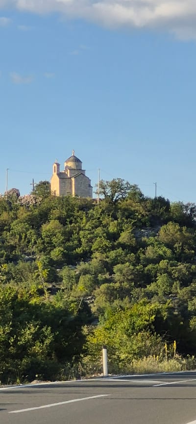 The image shows a view of the Church of Saint George in Liješnje, Montenegro, from a roadside perspective. The church, with its distinctive domes and structure, is situated on a hill or elevated terrain covered with dense greenery. A road with white lane markings is visible in the foreground, suggesting a nearby vantage point. The sky is blue with some clouds. Utility poles are visible running up the hill to the church.