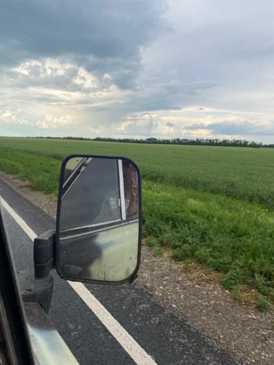 The image captures a perspective from inside a vehicle traveling along a paved road. A side view mirror is prominently featured, reflecting a person's head and shoulder. The road is bordered by a vibrant green field, and a distant treeline stretches across the horizon. Above, a dramatic sky filled with a mix of dark and light clouds suggests an approaching storm or changing weather. The scene appears to be an outdoor rural setting, consistent with the outskirts or countryside of Krylovskaya, Russia.