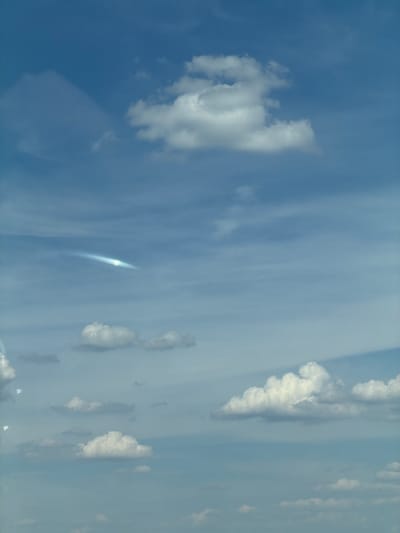 A sky scene with scattered white clouds against a bright blue sky. In the upper portion of the image, a large, fluffy cloud dominates the center. To the left of this cloud, a wispy, iridescent streak or contrail is visible, appearing as a faint, elongated glow. Further down, several smaller cumulus clouds are scattered across the frame. The overall impression is a bright, daytime sky, possibly taken from an airplane or a high vantage point. There are no discernable structures, people, or any indication of the city of Тэльмінскі сельскі Савет, Belarus.