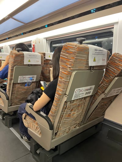 The image shows the interior of a train carriage, likely a high-speed train, with seats facing forward. The seats are upholstered in a patterned fabric with brown, orange, and white stripes. On the back of the headrests of the seats, there are white labels with text in Chinese and English, along with a yellow triangular warning symbol. One label visible in the foreground reads: "When adjusting the backrest, be careful not to tip over the tea cup behind you." The seats are designed with tray tables that fold down. A bottle of water is visible in the pocket of a seat. A woman with dark hair is seated in the foreground, facing away from the camera, and is holding a smartphone. Another person's head and shoulder are visible in the seat to the left. Above the seats, a digital display shows the temperature as 11 degrees Celsius, indicated by "11°C". The lighting in the carriage appears to be artificial, coming from overhead panels. The floor of the carriage is a dark grey material. This scene is set in the city of Longhua District, China.