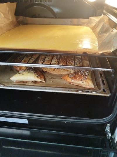The image shows the interior of an oven. On the top rack, a large, flat, yellow batter is being baked on parchment paper. On the middle rack, several triangular pastries with sesame and black seeds are arranged on a baking sheet, also lined with parchment paper. The oven is open, revealing the cooking food. There are no people or animals visible in the image. The environment is that of a kitchen oven, implying a home or commercial kitchen setting. There is no indication of the location being Andzhero-Sudzhensk, Russia.