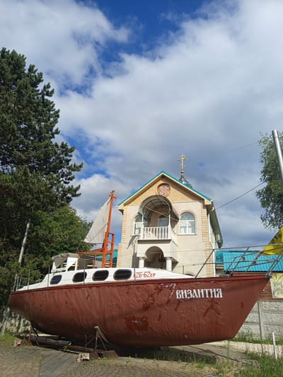 In the city of Giaginskaya, Russia, an old, partially weathered brown and white sailboat is displayed on a stand on a paved area. The sailboat is positioned in front of a tan, two-story building that appears to be a church, complete with a cross on its roof. The building features arched windows and a balcony. To the left of the sailboat, a large, green pine tree stands tall against a cloudy blue sky. The scene captures a static display with no people or activity evident.