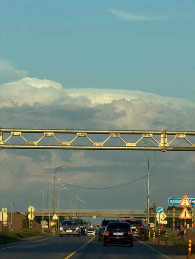 The image depicts a view from a vehicle traveling on a road, likely a highway, near Derzhavino, Russia. The road is paved with asphalt and has a yellow dividing line. Several cars are visible, moving in the same direction as the camera. On the right side of the road, there are multiple traffic signs, including directional signs with Russian text (e.g., "ЛАИШЕВКА" which likely refers to a location, and "СТОП" meaning "STOP"), as well as warning signs depicting a traffic light and a T-junction. There are also traffic cones and temporary barriers. Overhead, a large metal structure, possibly part of an overpass or a sign gantry, spans the road. In the background, there is an overpass or bridge structure. The sky is a mix of blue and gray clouds, with sunlight illuminating the clouds from the side, suggesting it is either early morning or late afternoon. The environment appears to be somewhat rural or suburban, with grassy areas and sparse vegetation visible on the sides of the road and beyond the barrier. The lighting creates strong shadows.