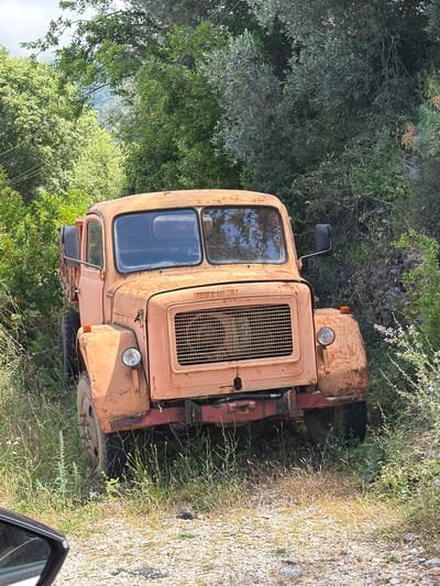 The image, captured in Bratešići, Montenegro, depicts an old, heavily rusted peach-orange colored vehicle, resembling a compact tractor or agricultural truck, parked amidst lush vegetation and a gravel path during the daytime.
The vehicle is the central subject, exhibiting significant signs of age and disuse, with widespread rust and faded paint covering its body, including the cabin, hood, and fenders. It features a cabin with two large, rectangular windows reflecting the bright sky and surrounding trees. A prominent, vertically oriented rectangular grille is visible on the front right side of the vehicle, with a single round headlight and an orange indicator light positioned nearby. A black side mirror extends from the left side of the cabin. The undercarriage and parts of the frame appear to be a darker, possibly reddish-brown color, contrasting slightly with the main body.
The vehicle is situated on a patch of dry grass and wildflowers, with dense green trees and bushes forming a verdant backdrop to its left. To the right, a light-colored gravel or dirt road/path is visible, also bordered by dry grass and sparse vegetation, including small purple flowers. The natural lighting suggests a sunny day with clear skies. No activity is taking place, and no people are visible, lending an atmosphere of abandonment and the quiet passage of time to the scene. No discernible text is visible on the vehicle or in the environment.