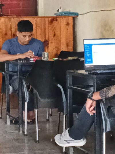 In the city of Meunasahtutong, Indonesia, a young man wearing a blue t-shirt is seated at a table, looking down at items on the table. He appears to be engaged in some activity with his hands, possibly arranging or handling small objects. To his right, another person's legs and arm are visible; this individual is wearing dark pants and a grey athletic shoe, and their arm is resting on the table, with a watch on their wrist. In the background, a wooden wall with vertical planks is visible, and a computer monitor is on a table to the right. The scene suggests an indoor setting, possibly a cafe or a study area.
