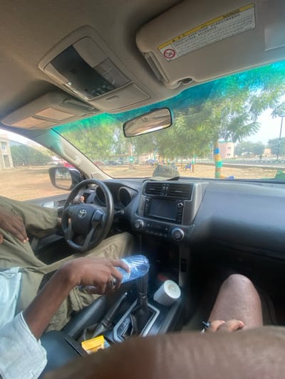 The image depicts an interior view from the front passenger seat of a Toyota Land Cruiser Prado SUV, parked outdoors in Yan Dala, Nigeria, during daylight hours.

In the foreground, the legs of the person occupying the passenger seat are visible, wearing shorts, with a dark object held in their right hand. The driver is seated to the left, dressed in a light-colored long-sleeved shirt and olive-green cargo pants. The driver's left hand, adorned with a silver-toned ring, holds a clear plastic water bottle above the center console. A small yellow box, possibly a matchbox, rests on the center console near the automatic gear shifter. A white roll of tape is placed in a cup holder or recess further along the console. A beige baseball cap with "LYNX" visible on its front is positioned on the dashboard directly in front of the passenger. The vehicle's dashboard features a touchscreen infotainment system, air vents, and climate controls. The passenger-side sun visor is raised, displaying a multi-language warning label for airbags.

Through the windshield, a dry, open area with numerous green-leafed trees is visible. In the background, a light-colored building is partially seen on the left. Towards the right, a multi-story building with distinct blue and pink/orange striped vertical elements is discernible. A paved road or pathway runs across the middle distance, and several other vehicles are parked further back, partially obscured by trees. Outdoor chairs are visible under some trees.