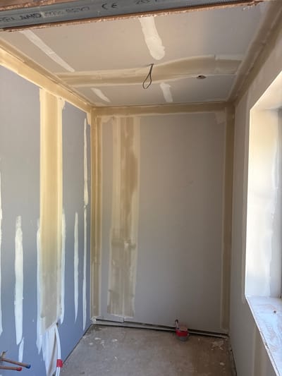 This media file depicts an unfinished interior room, likely a bathroom or utility space, in South Oxfordshire, United Kingdom, undergoing renovation or construction. The walls and floor are covered with plasterboard (drywall), and the surfaces are in the process of being prepared for painting.
Evidence of joint compound application is prominent, with white and beige streaks covering the seams and screw indentations on the plasterboard. On the right wall, a red plumbing fixture with exposed pipes suggests connections for water supply, while additional pipes with red and blue caps are visible on the adjacent floor area. A black electrical wire loop protrudes from a small hole in the left wall. The ceiling features a tiered bulkhead or dropped section, also finished with plasterboard and joint compound.
The scene captures an intermediate stage of interior finishing, specifically the taping and mudding phase of drywall installation. Lighting appears to be artificial. On a metal stud visible along the far left edge, partial text including "CE" and "BS EN" can be seen, indicating compliance with European and British standards, consistent with the specified location. The overall impression is of a functional room being readied for its final finishes.