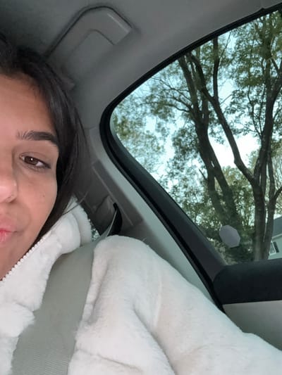 A female individual is partially visible in the left half of the image, seated in the front passenger seat of a vehicle in Lawrence Township, United States. She has dark hair and brown eyes, with her right eye, eyebrow, nose, and mouth visible. Her mouth is slightly pursed. She is wearing a white, fluffy, hooded jacket, zipped up, and a light-colored seatbelt is diagonally across her chest. The car's interior shows a gray headliner and a grab handle above the window. Through the vehicle's window, which occupies the right half of the frame, multiple deciduous trees with green foliage are visible against a light blue or overcast sky. A portion of a white house is discernible in the background, partially obscured by the trees. A small, light-colored, irregular sticker or reflection is present on the window glass, in front of the house. The scene appears to be static and takes place during daylight hours.