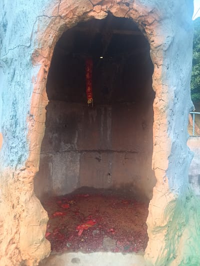 A small, artificial cave-like structure stands in Ban Thung, Thailand. The structure is painted to resemble rock and features a red banner with writing in an Asian script hanging inside. The floor of the "cave" is covered with red debris. The structure is open in the front, revealing its interior. The immediate surroundings include greenery and a partial view of a blue railing, suggesting an outdoor location.