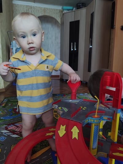 A young child with blonde hair and blue eyes stands in a room playing with a toy car garage. The child wears a yellow and gray striped polo shirt. The toy car garage is primarily red and gray with yellow accents. Part of another child's head is visible behind the toy. The room has a rug with a road pattern. In the background are a closet and a wall. The location is Malaya Purga, Russia.