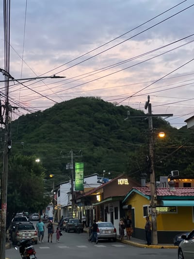 This is a street-level, eye-level, medium shot of a street in San Juan del Sur, Nicaragua. A large, lush green hill dominates the background, partially obscured by a dense network of power lines crisscrossing the sky. The sky itself is a pale pink and gray, indicating either dawn or dusk. The street is paved and lined with low-rise buildings, some painted in bright colors like yellow and teal. A few vehicles, including cars and a motorcycle, are parked or driving on the street. Several people are visible, walking on the sidewalk or crossing the street. One building has a neon sign that reads "HOTEL." Another building displays a green banner with "BANCO LAFISE BANCCENTRO." Streetlights are present and one appears to be illuminated. The overall impression is of a developing coastal town under a soft, atmospheric sky.