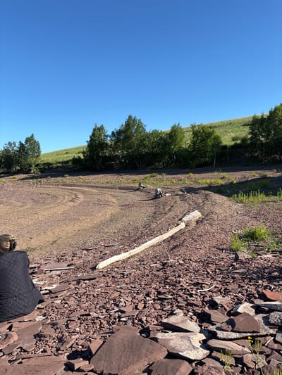 In Sovetskaya Khakasiya, Russia, a person is seated facing away from the camera in the foreground, dressed in a black quilted jacket. They are situated on a ground covered with numerous flat, reddish-brown stones and smaller gravel. A light-colored, elongated piece of wood rests horizontally in the mid-ground. The terrain is uneven, with mounds of reddish-brown material and sparse vegetation. In the background, a gentle green hillside rises, dotted with trees. The sky is a clear, bright blue.