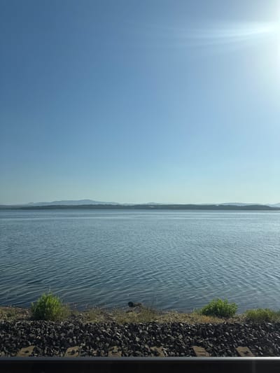 The image depicts a serene body of water, possibly a lake or a wide river, under a clear, bright sky. The sun is high, indicated by the strong, diffused light and lens flare in the upper left portion of the image. The water's surface is rippled, suggesting a gentle breeze.
On the right side of the frame, the water meets a rocky shoreline covered with sparse green vegetation. The rocks appear to be a mix of dark, coarse stones, with some larger, lighter-colored rectangular objects, possibly concrete blocks, interspersed among them. A dark, vertical structure, possibly a railing or part of a bridge, is visible along the far right edge.
In the distance, across the water, a tree-lined shore is visible, with hills or mountains forming a hazy, layered silhouette against the sky. The overall scene conveys a sense of tranquility and natural beauty, likely captured during the daytime, possibly late morning or early afternoon, given the brightness of the sun. There is no visible text in the image. The location context provided, Bages, France, suggests a riparian or coastal setting.