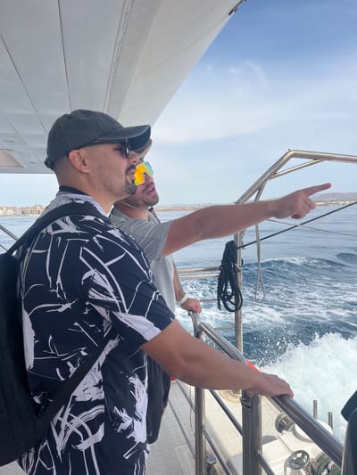 Two men are on a boat with the sea in the background. The man in the foreground is wearing a dark baseball cap, sunglasses, and a black and white patterned short-sleeved shirt. He has a black backpack on his left shoulder. The man behind him is wearing bright, multi-colored sunglasses and a grey t-shirt. He is pointing his right index finger towards the right of the frame. The boat has metal railings and the sea is choppy with white foam. In the distance, a coastline with buildings is visible, suggesting the location is near a city. Given the presence of a coastline with buildings and the sea, this scene could potentially be in Elx/Elche, Spain, which has a coastal area.