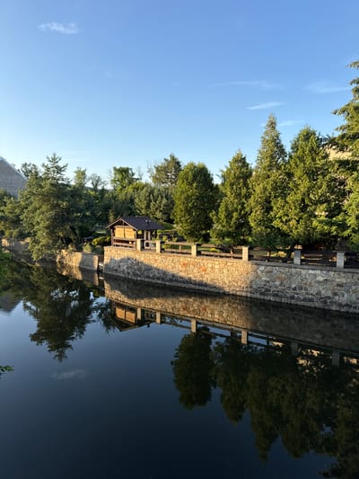 This image depicts a tranquil waterside scene in Beroun, Czechia, likely during late afternoon or early evening, given the warm lighting and long shadows. A dark, reflective body of water, possibly a canal or river, occupies the lower portion of the frame. The water's surface is calm, mirroring the stone wall and vegetation lining its bank.
The bank itself is reinforced with a substantial, textured stone wall constructed from irregularly shaped, light-brown and grey stones. Above the stone wall, a wooden fence with upright supports adds a rustic charm to the scene. Behind the fence, a lush green area is populated by a variety of trees, predominantly evergreen, with some deciduous trees interspersed.
Prominently featured on the elevated bank is a small, wooden structure with a dark, pitched roof. It appears to be a pavilion or a small shelter, with open sides supported by wooden posts. The structure has a rustic, almost quaint appearance, with visible wooden planks forming its walls. The area around the structure is well-maintained, with manicured bushes and more trees creating a green backdrop.
To the left of the pavilion, a larger, modern building with a grey, angled roof is partially visible, suggesting a contemporary architectural presence in contrast to the older, natural elements.
The sky above is a clear, vibrant blue with a few wispy clouds. The sunlight illuminates the stone wall, casting a warm glow and creating distinct shadows, highlighting the textures and depth of the scene. The reflections in the water are sharp and clear, doubling the visual impact of the architecture and the surrounding greenery. There are no people visible in the image, contributing to the overall sense of peace and quiet. The scene evokes a feeling of serenity and natural beauty, possibly a public park or a private garden area within the city of Beroun.