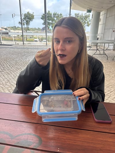 A young woman with long, straight, light brown hair is seated at a reddish-brown wooden table, looking directly at the camera. She is wearing a dark top or jacket, small gold hoop earrings, and a delicate gold necklace. Her left hand holds a silver spoon, positioned near her open mouth, suggesting she is eating. A gold ring is on her left ring finger, and her fingernails are painted dark red. On the table in front of her is a rectangular clear plastic food container with a blue lid, partially open, containing what appears to be a mixed dish. To the right of the container, a smartphone in a purple case rests on the table. The background features large glass windows overlooking a paved outdoor area with a patterned surface. Beyond, green trees, distant residential buildings on hills, and several white vehicles are visible under a partly cloudy sky. Inside, behind the woman, several light-colored tables with metal frames and a white cylindrical column are present. A green exit sign is mounted overhead to the right. The scene is located in Porto Salvo, Portugal.