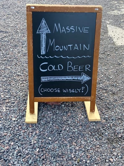 A wooden A-frame chalkboard sign stands on a gravel surface. The chalkboard is black with white chalk writing and drawings. At the top, a hand-drawn arrow points upward. Below the arrow, the words "MASSIVE MOUNTAIN" are written in block letters. Beneath that, "COLD BEER" is written in large, bold letters. To the right of "COLD BEER" is a hand-drawn arrow pointing to the right. Below the arrow, enclosed in parentheses, is the text "(CHOOSE WISELY!)". The sign is positioned outdoors on a surface covered with small, gray, and reddish-brown gravel. The background is blurred and appears to be more of the same gravel. The location is Nikkaluokta, Sweden.