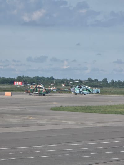 The image shows two helicopters on the tarmac at Khrabrovo Airport in Khrabrovo, Russia. The helicopter on the left is a camouflage-patterned military helicopter with a red star insignia. It is parked on the tarmac with its blades extended. The helicopter on the right is a lighter-colored helicopter, possibly a civilian or different military model, also parked on the tarmac. In the background, there are trees and buildings, including what appear to be airport structures. The sky is cloudy. The tarmac is marked with various painted lines. An orange marker cone is visible to the left of the military helicopter. The overall scene depicts an active or recently active airfield.