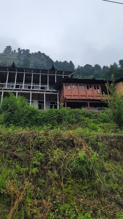 The image captures a view of traditional buildings nestled on a hillside in Cheelaage, India. The architecture features multi-story structures with prominent wooden balconies and roofs, possibly indicative of a mountain or hilly region. The buildings are surrounded by dense green foliage and vegetation, with the hillside showing signs of natural growth. The sky is overcast and appears cloudy or foggy, suggesting a humid or temperate climate. The scene depicts a rural or semi-urban setting, emphasizing the integration of human habitation with the natural landscape.