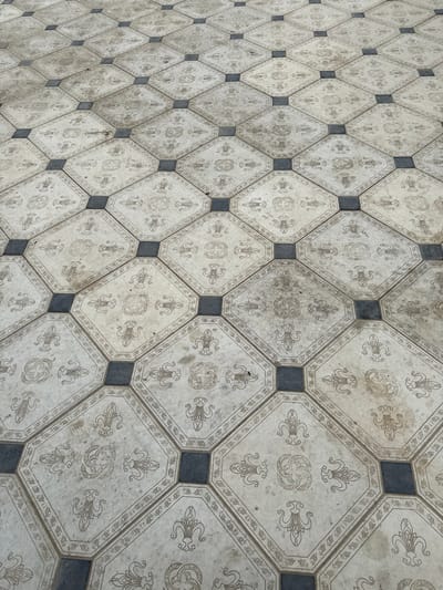 The image shows a close-up, eye-level view of an outdoor tiled floor. The tiles are arranged in a repeating pattern of octagons with decorative designs and small square inserts in dark gray. The octagonal tiles are light beige with intricate, repeated patterns in a darker brown or gray, including what appear to be stylized floral motifs and circular designs. The overall impression is of an aged or weathered surface, with some areas of discoloration and a visible layer of dust or dirt. The perspective is from above, looking down at an angle, and the floor extends into the distance, creating a sense of depth. There are no people or specific landmarks visible. The image was taken in the city of Beshtal, Uzbekistan.