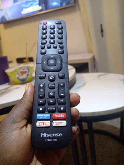 The image showcases a Hisense TV remote control, model EN2BI27H, held in a hand. The remote features standard buttons like numbers, volume, channel control, directional buttons, and 'OK.' It includes dedicated buttons for quick access to streaming services: Prime Video, YouTube, Netflix, and Rakuten. The remote is black with white text and colored logos for the streaming apps. The background is blurred, showing elements of a room with a TV visible at the top.