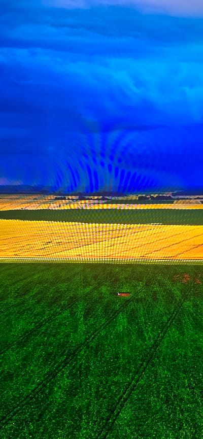 This is an aerial photograph of agricultural fields near Lippstadt, Germany, under a dramatic, dark blue sky. The image is divided into three horizontal bands of color. The top band is a deep, textured blue representing the sky and clouds. The middle band is a vibrant yellow, likely a wheat or barley field, exhibiting horizontal rows and some texture. A thin, horizontal line of discolored pixels runs through this yellow band. The bottom band is a rich, dark green, representing another field, possibly grass or a different crop, with visible tire tracks from agricultural machinery. In the lower right portion of the green field, a small, rectangular, reddish-brown object, possibly a piece of equipment or debris, is present. The overall impression is one of vast, open farmland under a foreboding sky.