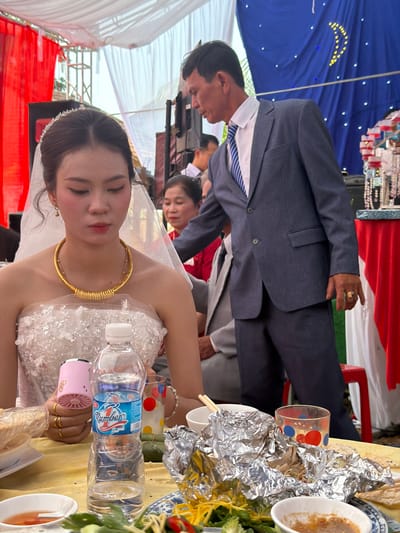 This image captures a daytime celebratory event, likely a wedding or engagement ceremony, set outdoors under a large tent or canopy. The scene is illuminated by bright, diffused light.
The primary subjects are a woman, presumably the bride, and a man, likely the groom. The bride, positioned in the foreground, wears a white, strapless or off-the-shoulder gown adorned with sequins or beads. She accessorizes with a large, golden choker necklace, a small jeweled tiara or headband, and gold rings on her right hand. She holds a small pink portable fan and a plastic water bottle. Her expression is serene. The groom, standing behind the bride, is dressed in a dark blue suit, a white collared shirt, and a blue and white striped tie. He wears a gold ring on his left hand.
Other individuals are visible in the background, including a woman in a red top and several partially obscured faces, suggesting a gathering of guests.
The table in the foreground is covered with a yellow tablecloth and features various dishes, some wrapped in aluminum foil, bowls, chopsticks, and a clear glass with red and blue polka dots. A plastic water bottle with a blue label, where the brand "Jumboz" (or similar) is partially visible, is also on the table.
The setting includes blue fabric draped overhead, decorated with yellow and white star-like embellishments, possibly forming a crescent moon. White canopy fabric is visible, along with red fabric draping in the background. A jewelry display stand with necklaces and bracelets is faintly seen in the far background.
Given the context of Tay Son Commune, Vietnam, the attire, decorations, and presence of specific food items (like a green wrapped item on the table, potentially a traditional cake) are consistent with a Vietnamese wedding celebration.