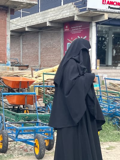 In Gulab Bagh, India, a person wearing a black niqab is standing in an outdoor setting. The person is positioned to the right of the frame, facing towards the left. In the foreground, there are several orange wheelbarrows stacked on blue metal frames, with yellow wheels. Behind the wheelbarrows, there are piles of wood and some green metal carts. In the background, a building with brick walls and a concrete overhang is visible. A sign with the Anchor logo is mounted on the building. The ground is covered with gravel and some patchy grass. The lighting suggests it is daytime.