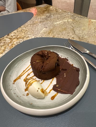 A chocolate lava cake with a scoop of vanilla ice cream and a slice of chocolate garnish rests on a white, grey-rimmed plate. The dessert is drizzled with caramel sauce and sprinkled with gold flakes. The plate sits on a dark grey placemat atop a granite countertop. A fork and knife are visible to the right of the plate. The setting appears to be a restaurant in Shkotovo, Russia, with a blurred background of people and furniture.