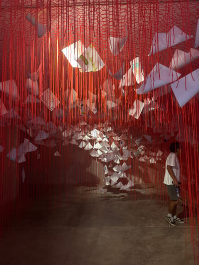 This image captures an indoor installation in San Isidro Llano Grande, Mexico. Thousands of red strings descend vertically from a red net-like ceiling structure, forming a dense curtain that fills the space. Multiple white papers, some folded or creased and bearing faint markings or drawings, are suspended at varying heights within the string curtain. Overhead illumination highlights the papers, causing them to glow brightly against the dim background. The floor is a dark, smooth surface. On the right side of the frame, one male individual stands in profile, facing left, observing the illuminated papers. He is dressed in a white short-sleeved t-shirt, dark shorts, and black and white athletic shoes. He holds a dark-colored bottle in his right hand. The immediate environment is dimly lit, with focused illumination directed at the suspended papers and upper string layers.